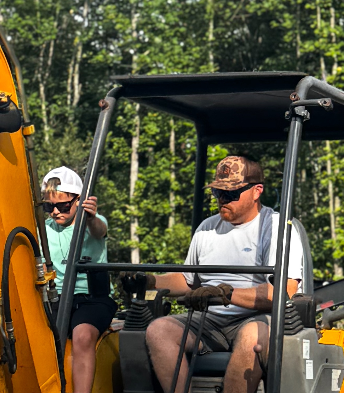 Father and child in an excavator cab, dad wearing the Evergreene Excavation bark camo hat